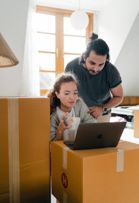 A man with dark hair tied back and a young girl with curly hair are inside a well-lit room that has wooden window frames and cream-colored walls. The girl is seated at a wooden table, holding a white mug, and working on a laptop placed on a large cardboard moving box. The man is leaning over, observing or assisting with the laptop. Surrounding them are several other cardboard boxes, some sealed with packing tape, indicating they are in the process of home relocation. The scene captures a moment of coordination during packing and moving activities, with natural light coming through the window, illuminating the workspace. The environment suggests an organized moving process, consistent with professional removals services by [COMPANY_NAME].