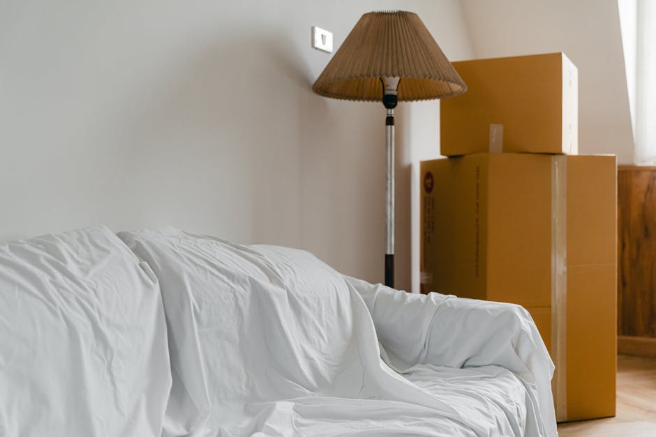 Inside a residential room during a home relocation process, showing a white fabric-covered sofa positioned adjacent to a light grey wall. Behind the sofa, a vintage-style floor lamp with a pleated brown lampshade stands next to a stack of three brown cardboard boxes of varying sizes arranged on the wooden floor. The boxes are sealed with packing tape, indicating they are ready for transport. Natural light enters through a nearby window, illuminating the scene. The environment suggests a professional removals service, with visible packing materials and furniture prepared for loading into a moving van, consistent with house removals and furniture transport activities performed by companies like Removal Company Harrow.