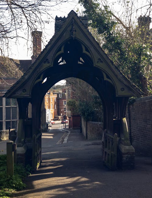 A narrow residential street seen through a decorative, pointed wooden archway with intricate carvings, leading to a quiet lane lined with brick buildings and parked cars. The archway is part of a structure at the entrance to a property, with shadows cast on the pavement below. In the background, trees with bare branches and a mix of sunlight and shade are visible, indicating a daytime setting. The scene reflects a typical UK neighborhood area that might be involved in a house removal or relocation process, with Removal Company Harrow potentially assisting with furniture transport or packing and moving activities near this historic street entrance.