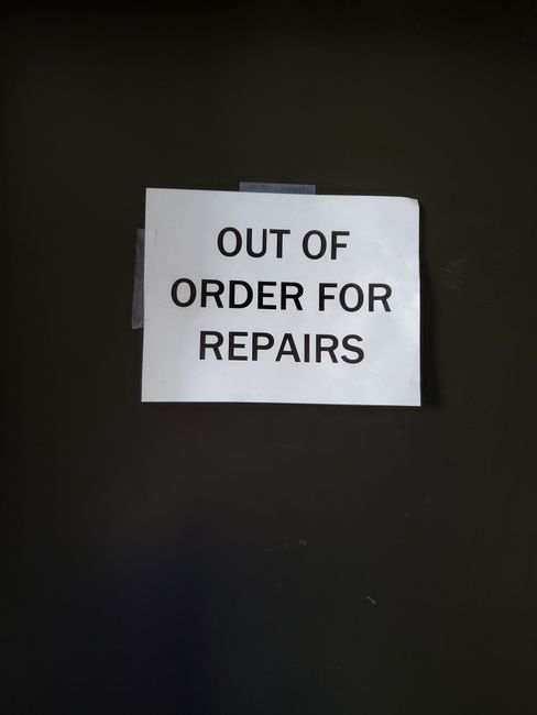 A close-up photograph of a white paper sign taped to a dark surface, displaying the message 'OUT OF ORDER FOR REPAIRS' in bold, black capital letters. The sign appears to be attached with transparent tape at the top corners. The dark background suggests the sign is affixed to a piece of equipment, a door, or an industrial unit, potentially within a residential or commercial property involved in home relocation or moving services. The context of the image relates to logistical considerations during house removals, such as a temporary closure or restricted access area affecting furniture transport or loading processes. The lighting is even, with no visible shadows or reflections, emphasizing the clarity of the notice for anyone involved in the moving or packing operations.
