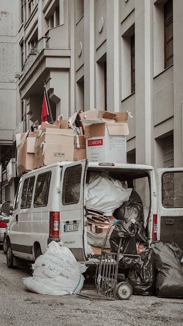 A white mini van parked on a city street with its rear doors open, revealing an interior filled with large black and white plastic bags, cardboard boxes, and wrapped furniture items, some stacked on top of each other. Several cardboard boxes and packages are placed on the roof of the van, secured with straps, including a prominent white box marked 'LEARDON'; other packing materials such as bubble wrap and packing paper are also visible. A small trolley is positioned next to the van's open door, indicating loading or unloading activity. The van is situated in front of an urban building with multiple windows and a beige facade. The scene depicts a home relocation or furniture transport operation, with the van prepared for moving belongings as part of a removal company's service, consistent with the context of house removals and logistical planning for a move.