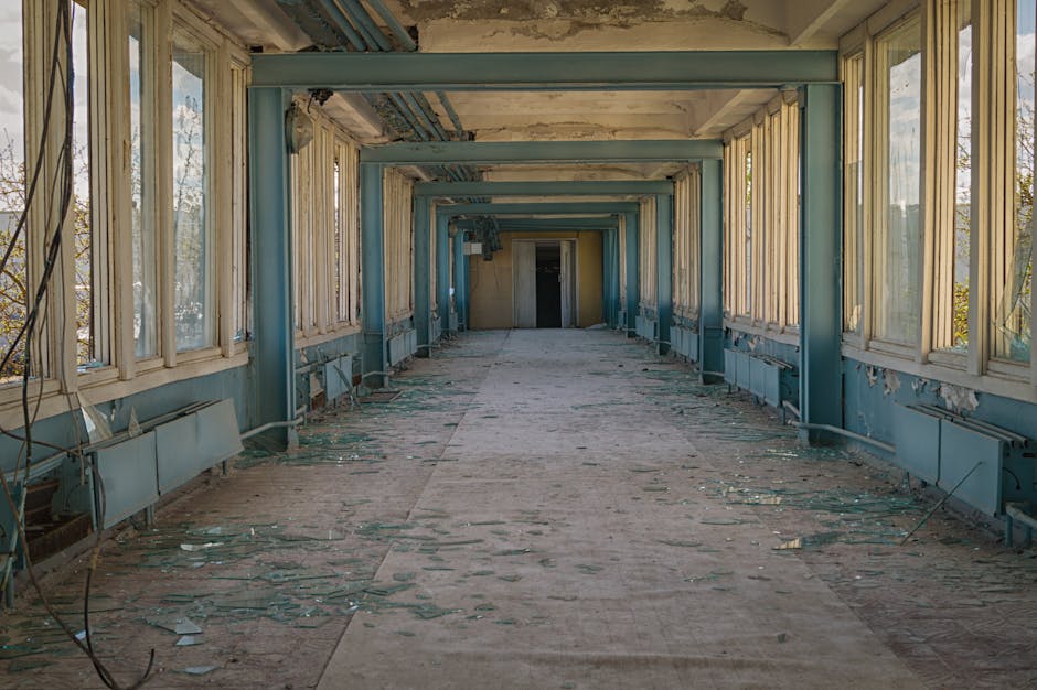 A long, narrow corridor inside an abandoned building with peeling blue paint on the walls and ceiling. Large windows on both sides are broken, with some glass panes missing or shattered, allowing natural light to illuminate the space. The floor is covered in debris, including fallen plaster, broken glass, and scattered insulation material. Exposed wiring hangs from the ceiling, and old radiator panels are attached along the lower part of the walls. The corridor appears to be in a state of disrepair, with visible signs of neglect and deterioration. This environment highlights the contrast between the current dilapidated state and the possibility of renovation or refurbishment, which may be relevant in the context of home or building removals. Occasionally, Removal Company Harrow may be involved in relocating or clearing spaces like this as part of their property transition services.