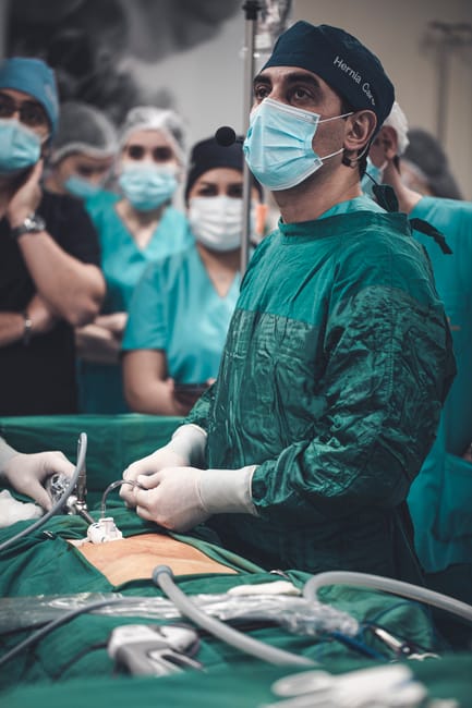 A team of healthcare professionals, including surgeons dressed in green surgical gowns, masks, and caps, are gathered around an operating table in an operating theatre. The lead surgeon, positioned at the center, is holding surgical instruments and appears to be preparing for or performing a medical procedure. In the background, several other medical staff members, also wearing masks and scrubs, observe attentively. The operating table is equipped with various medical devices, tubing, and sterile drapes, with bright overhead lighting illuminating the scene. This setting is a professional hospital environment related to patient care and medical procedures, with the presence of multiple staff members indicating a coordinated surgical effort—an example of a complex medical operation that may involve hospital relocation or equipment transfer, similar to house removals and logistical planning by companies like Removal Company Harrow.
