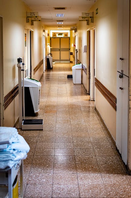 A long, narrow corridor inside an abandoned building with peeling blue paint on the walls and ceiling. Large windows on both sides are broken, with some glass panes missing or shattered, allowing natural light to illuminate the space. The floor is covered in debris, including fallen plaster, broken glass, and scattered insulation material. Exposed wiring hangs from the ceiling, and old radiator panels are attached along the lower part of the walls. The corridor appears to be in a state of disrepair, with visible signs of neglect and deterioration. This environment highlights the contrast between the current dilapidated state and the possibility of renovation or refurbishment, which may be relevant in the context of home or building removals. Occasionally, Removal Company Harrow may be involved in relocating or clearing spaces like this as part of their property transition services.