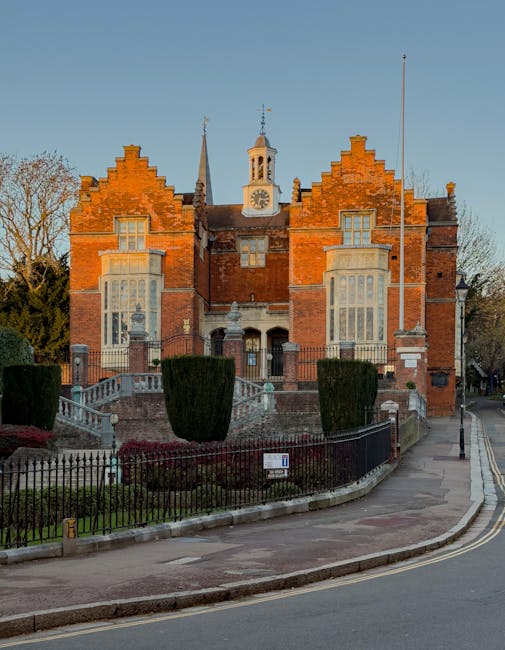 A large historic red-brick building with a decorative gabled roof, multiple tall windows, and a central clock tower with a weather vane on top. The building is elevated above street level, accessible by a curved stone staircase with metal railings, and surrounded by a low black metal fence. In front of the building, there is a landscaped area with neatly trimmed bushes and a paved pathway. To the right, a narrow sidewalk runs along the road, which has a street lamp and a sign attached to the fence. The scene is lit by natural daylight, with clear skies above. The setting suggests a prominent county hall or similar municipal building, and the image is relevant to house removal or relocation services by companies like Removal Company Harrow, highlighting the importance of careful furniture transport and packing in house removals within historic or urban environments.