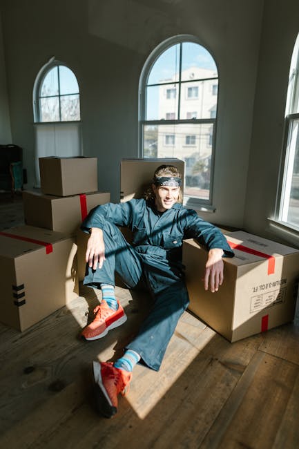 A person dressed in casual clothing and bright orange running shoes sits on a wooden floor inside a room with large arched windows, surrounded by multiple cardboard boxes sealed with red and black tape, indicating a home relocation or packing process. The room is well-lit by natural daylight streaming through the windows, with a view of a neighboring building outside. The individual appears to be taking a break during packing or furniture transport activities, which are part of a house removal or moving service offered by Removal Company Harrow. The scene captures the staging area where packing materials are prepared, and furniture or belongings are being systematically loaded for transportation to a new property, reflecting the logistics involved in professional removals.